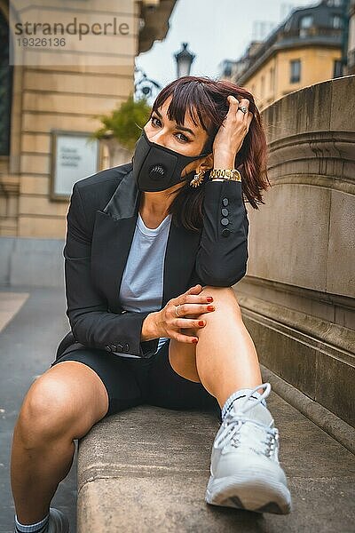 Modefotoshooting der neuen Normalität  eine junge Brünette mit einer Maske auf dem Gesicht von der Coronaviruspandemie sitzend in der Stadt. Mangelndes Vertrauen nach dem Covid19 soziale Distanz