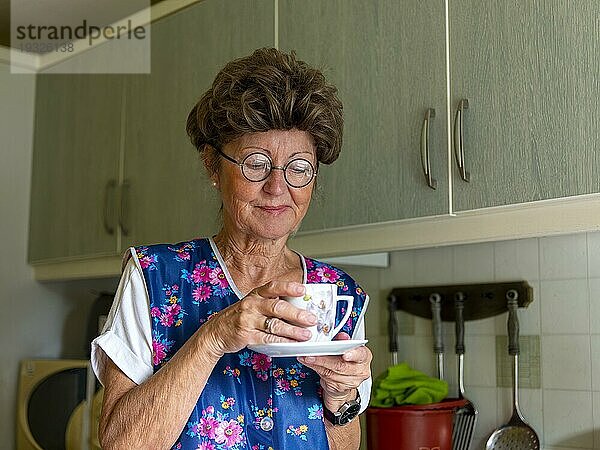 Oma mit alter Kittelschürze  Brille und Perücke in der Küche  hält Kaffeetasse in Händen  Deutschland  Europa
