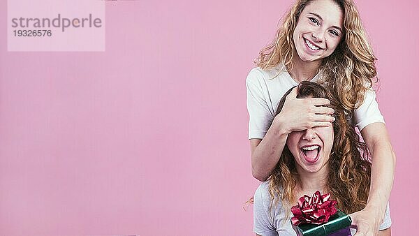 Frau bedeckt die Augen ihres Freundes und gibt ihm eine Geschenkbox vor einem rosa Hintergrund