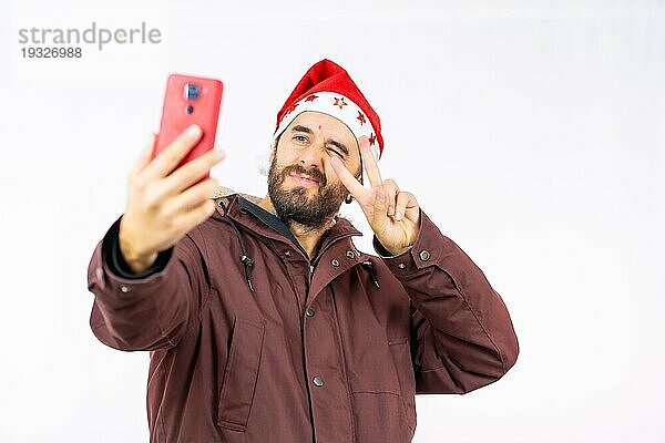 Sehr glücklicher junger kaukasischer Mann mit roter Weihnachtsmütze  der ein Selfie auf weißem Hintergrund macht  bekleidet mit einer Winterjacke