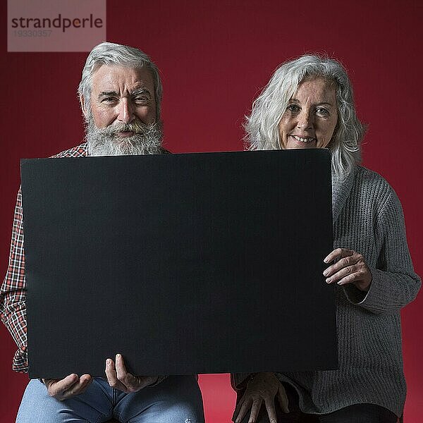 Porträt glückliches älteres Paar  das ein schwarzes Plakat hält und in die Kamera schaut  vor rotem Hintergrund