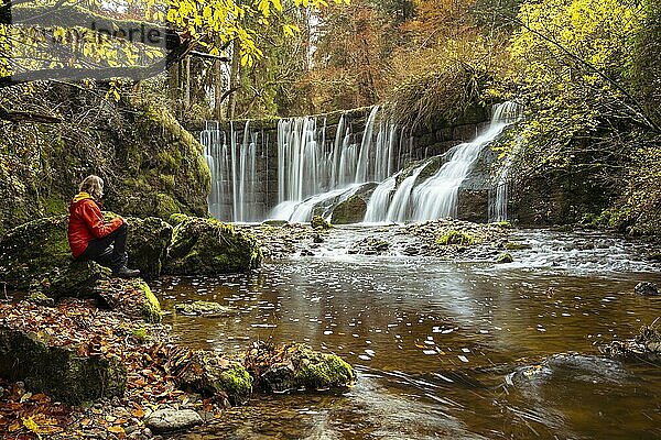 Der Geratser Wasserfall im Herbst. Eine Wanderin sitzt auf einem Felsen. Moosbedeckte Felsen an den Seiten  Bäume im Herbstlaub. Gerats  Oberallgäu  Allgäu  Bayern  Deutschland  Europa