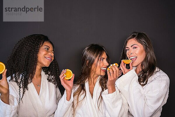 Studiofoto mit grauem Hintergrund von multiethnischen entspannten Frauen im Bademantel  die Obst essen
