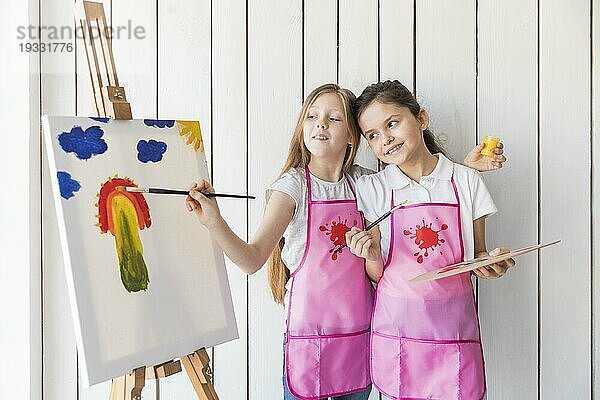 Glückliches Mädchen hält Palette Hand suchen ihre Freundin Malerei Leinwand mit Pinsel