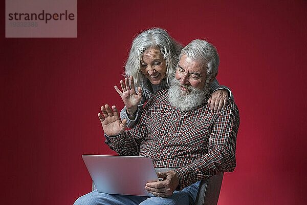 Älteres Paar schaut auf den Laptop und winkt mit den Händen vor rotem Hintergrund
