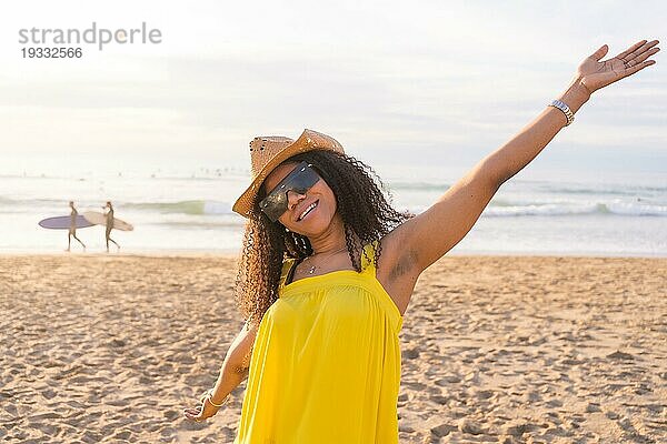 Gemischtrassige glückliche Frau mit Sonnenbrille und Sonnenhut  die die Arme hebt und Wohlbefinden am Strand signalisiert