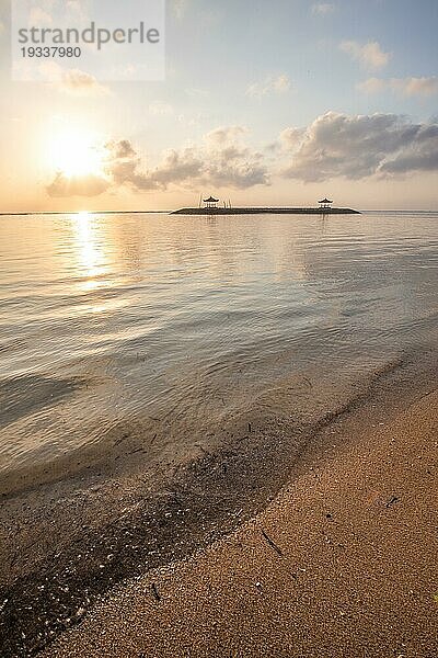 Sonnenaufgang über dem Meer. Blick vom Sandstrand bis zum Horizont. Im Meer gibt es Wellenbrecher mit kleinen Tempeln. Ruhiges Wasser mit kleinen Wellen und Reflexionen am Tropischen Strand von Sanur  Bali  Indonesien  Asien