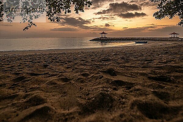 Sonnenaufgang über dem Meer. Blick vom Sandstrand bis zum Horizont. Im Meer gibt es Wellenbrecher mit kleinen Tempeln. Ruhiges Wasser mit kleinen Wellen und Reflexionen am Tropischen Strand von Sanur  Bali  Indonesien  Asien