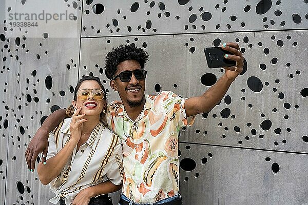 Multiethnische Freunde  die sich umarmen  während sie ein Selfie mit einem Mobiltelefon vor einem städtischen Hintergrund machen