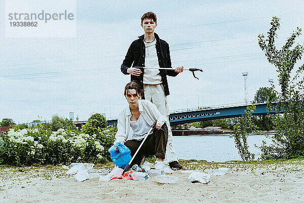 Ganzkörperporträt selbstbewusster junger Freunde  die Plastikmüll in der Nähe eines Flusses vor dem Himmel sammeln