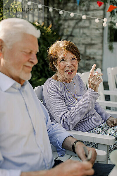 Porträt einer lächelnden älteren Frau  die gestikuliert  während sie neben einem Mann im Hinterhof sitzt