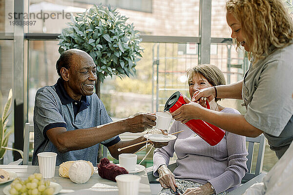 Junge Krankenschwester serviert einem pensionierten älteren Mann  der neben einer Frau im Pflegeheim sitzt  Kaffee
