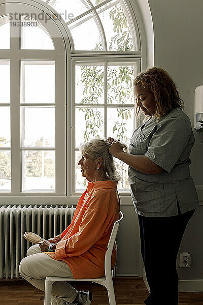 Seitenansicht einer jungen Pflegerin  die einer Seniorin im Pflegeheim die Haare flechtet
