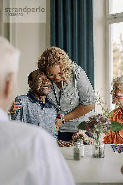 Lächelnde junge Pflegerin umarmt glückliche ältere Männer  die mit Freunden am Esstisch im Pflegeheim sitzen