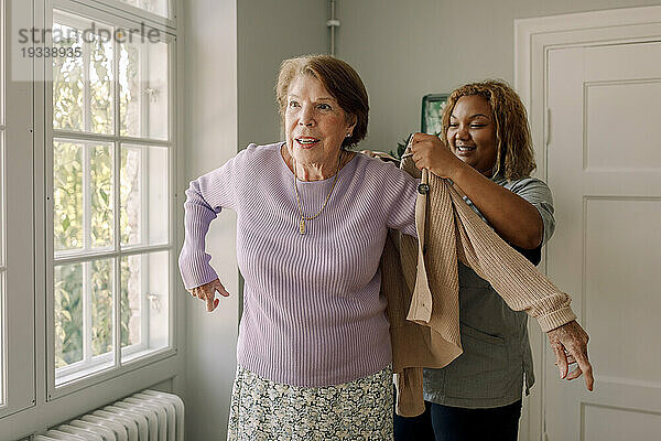 Lächelnde Krankenschwester hilft einer älteren Frau beim Tragen eines Pullovers im Altersheim
