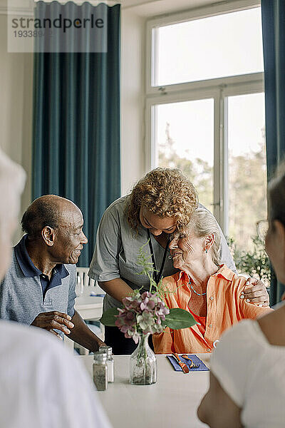 Junge Pflegerin umarmt ältere Frau  die mit Freunden am Esstisch im Pflegeheim sitzt