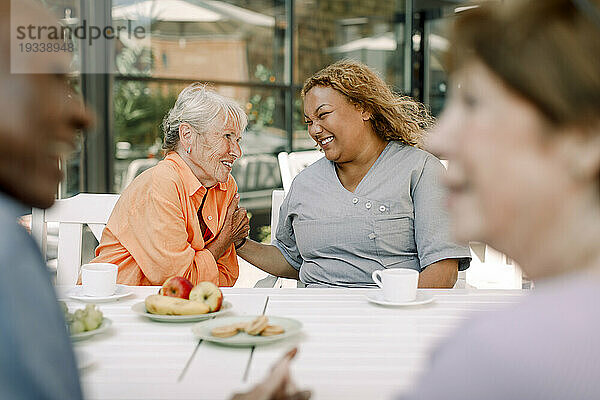 Fröhliche junge Betreuerin sitzt mit älteren Männern und Frauen im Ruhestand am Esstisch