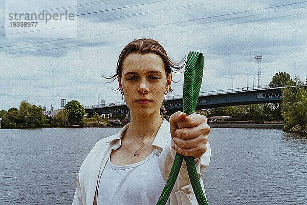 Porträt einer selbstbewussten jungen nicht-binären Person  die einen Gartenschlauch in der Nähe eines Flusses hält