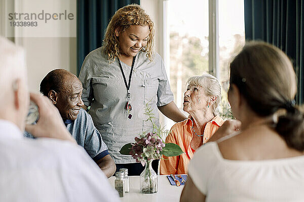 Lächelnde junge Pflegerin im Gespräch mit älteren Frauen und Männern am Esstisch im Pflegeheim