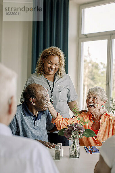 Lächelnde junge Pflegerin steht neben glücklichen älteren Frauen und Männern am Esstisch im Pflegeheim