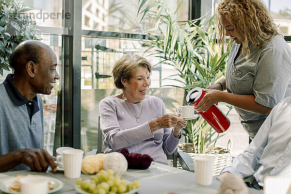 Junge Krankenschwester serviert einer pensionierten Seniorin  die neben einem Mann im Pflegeheim sitzt  Kaffee
