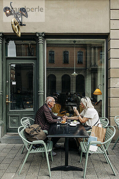 Ganzkörperansicht eines älteren Mannes  der eine Frau an der Hand hält  während er zusammen im Straßencafé sitzt