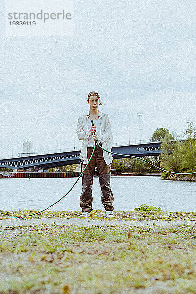 Ganzkörperporträt einer selbstbewussten jungen nicht-binären Person  die einen Gartenschlauch in der Nähe eines Flusses hält