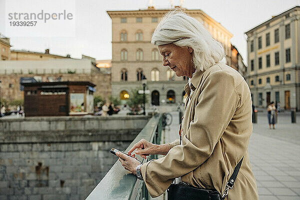 Seitenansicht einer älteren Frau  die sich auf ein Geländer lehnt  während sie in der Stadt ihr Smartphone benutzt