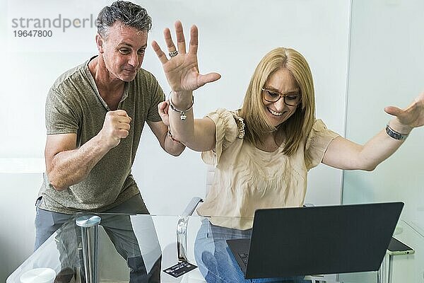 Fröhliches Ehepaar kauft von zu Hause aus mit Notebook und Kreditkarte ein und hebt dabei die Arme hoch