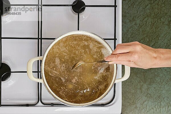 Draufsicht auf eine nicht erkennbare Frau  die eine Suppe in einer Kasserolle auf einem Gasherd anrührt