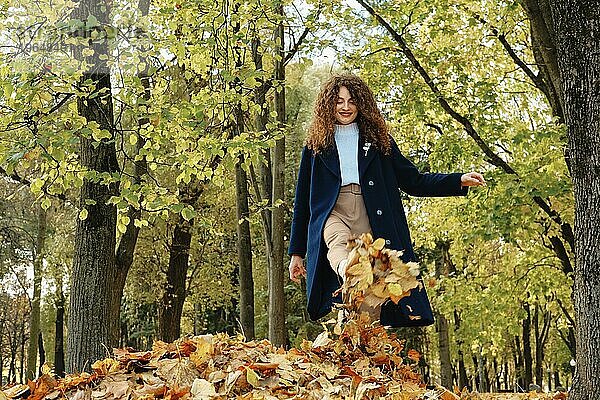 Niedriger Blickwinkel auf junge glückliche Frau  die Herbstblätter im Park aufwirbelt und lacht