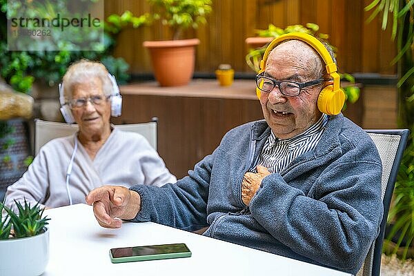 Glücklicher alter Mann  der mit Kopfhörern in einer geriatrischen Einrichtung neben anderen älteren Menschen Musik hört