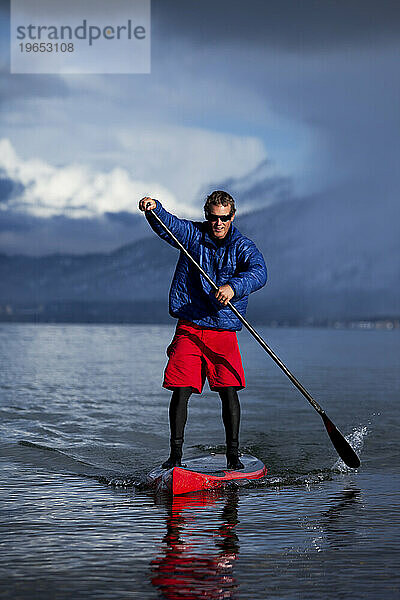Ein Mann beim Stand-Up-Paddleboarden auf einem Bergsee im Winter  während ein Sturm aufzieht.