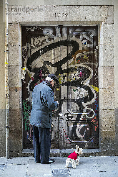 Älterer Herr mit kleinem Hund im roten Pullover an der Leine hält inne  um seine mit Graffiti bedeckte gotische Tür in Barcelona aufzuschließen.