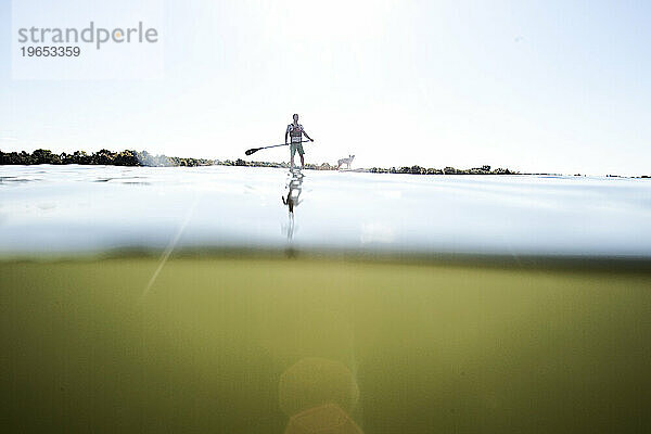 Split-Level-Bild eines Mannes beim Stand-Up-Paddleboarden mit seinem Hund vor einem verwehten Himmel.