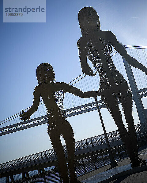Metallskulptur von zwei Personen mit einer Brücke im Hintergrund.