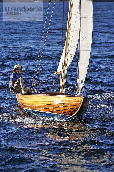 Ein fröhlicher Teenager fliegt durch das Wasser und segelt in einem klassischen Holzboot in der späten Nachmittagssonne.