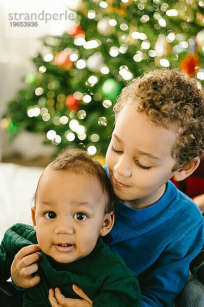 Gemischtrassiges Kind hält seinen kleinen Bruder vor dem Weihnachtsbaum