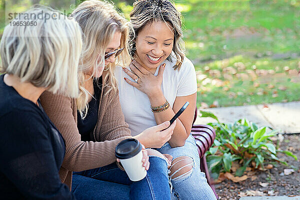 Drei Frauen sitzen auf einer Bank und lachen über das  was sie auf einem Telefon sehen