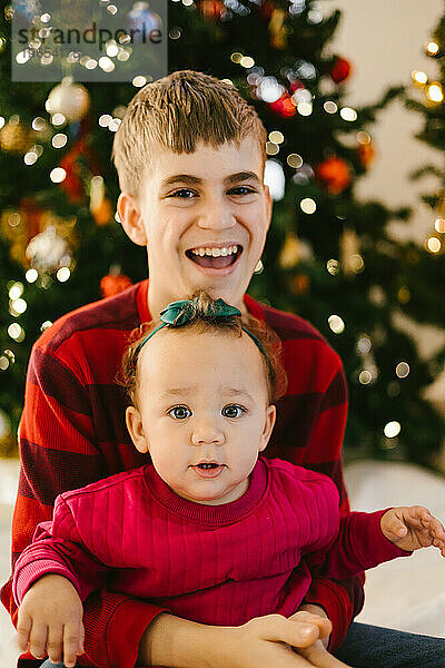 Bruder hält seine kleine Schwester lächelnd vor dem Weihnachtsbaum auf dem Schoß