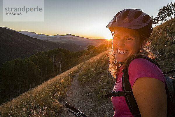Eine Frau auf ihrem Mountainbike mit Helm blickt lächelnd in die Kamera bei Sonnenuntergang.