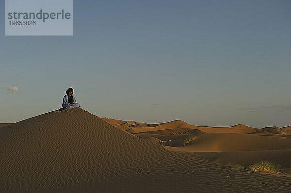 Ein Wüstenführer in traditioneller Kleidung sitzt bei Sonnenuntergang auf einer Düne.