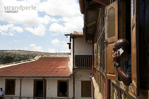 Ein äthiopischer junger Mann lehnt sich aus dem Fenster eines alten Hauses mit Blick auf die Stadt  während er mit seinem Mobiltelefon telefoniert.
