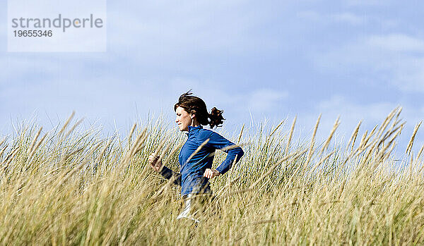 Eine Frau läuft einen Sandweg mit hohem Gras am Meer entlang.