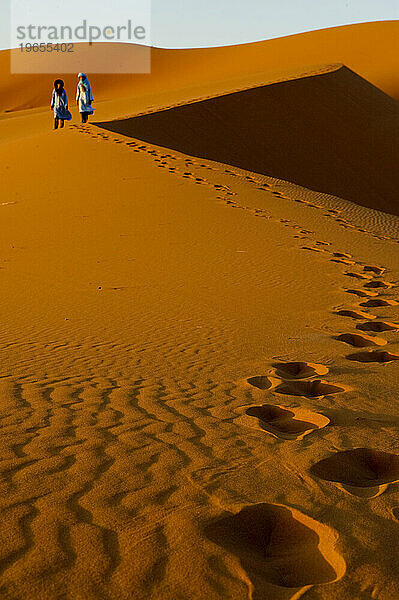 Two boys in traditional North African dress stand on the top of a sand dune at sunrise.
