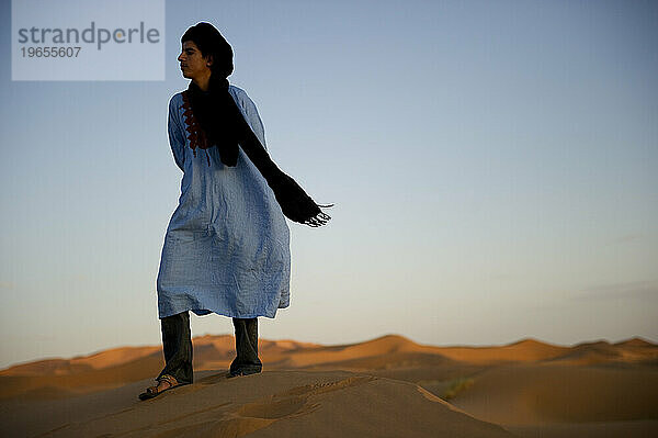 A desert guide in traditional dress stands on a dune at sunset.