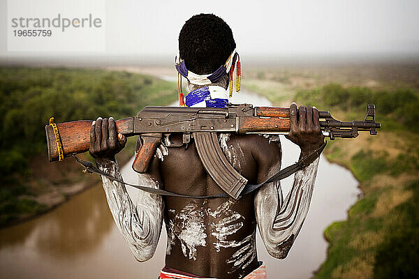 Ein junger Mann hält sein Kalaschnikow-Gewehr in der Hand  während er den Omo-Fluss im abgelegenen Omo-Tal in Äthiopien überblickt
