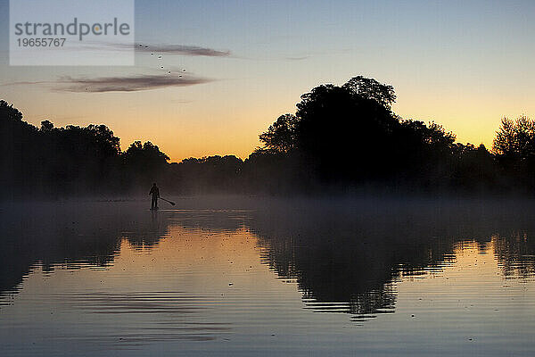 Silhouette eines Mannes beim Stand-Up-Paddleboarden im Sonnenaufgangslicht mit Bäumen  die sich im Wasser spiegeln.