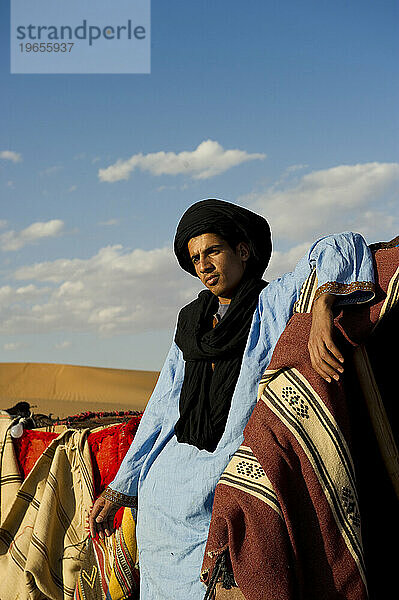 Ein Wüstenführer in traditioneller Kleidung steht in einem Zeltlager mit bunten Decken.