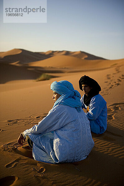 Zwei Jungen in traditioneller nordafrikanischer Kleidung sitzen bei Sonnenaufgang auf einer Sanddüne.